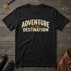 A black short-sleeve graphic t-shirt laid flat on a dark wood background, featuring the text 'ADVENTURE IS THE DESTINATION' in a distressed cream and orange font. The shirt is styled with a key, brown leather shoes, and a brown leather jacket around it.