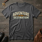 A dark heather gray t-shirt with a vintage-style graphic reading 'ADVENTURE IS THE DESTINATION' in cream and orange lettering, laid flat on a rustic dark wood background with keys, shoes, and a leather jacket as props.
