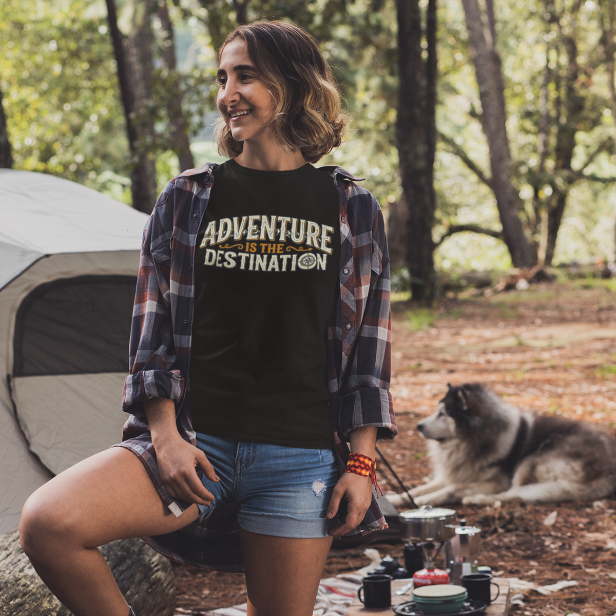 A woman posing in a forest campsite setting, wearing a black graphic t-shirt with the text 'ADVENTURE IS THE DESTINATION' in yellow and white, layered under an open plaid flannel shirt. A gray tent is visible on the left, and a dog is lying on the ground in the background.