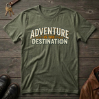 An military green graphic t-shirt laid flat on a dark wood background, featuring the text 'ADVENTURE IS THE DESTINATION' in cream and orange. The scene is styled with a leather jacket, keys, and a pair of black shoes.
