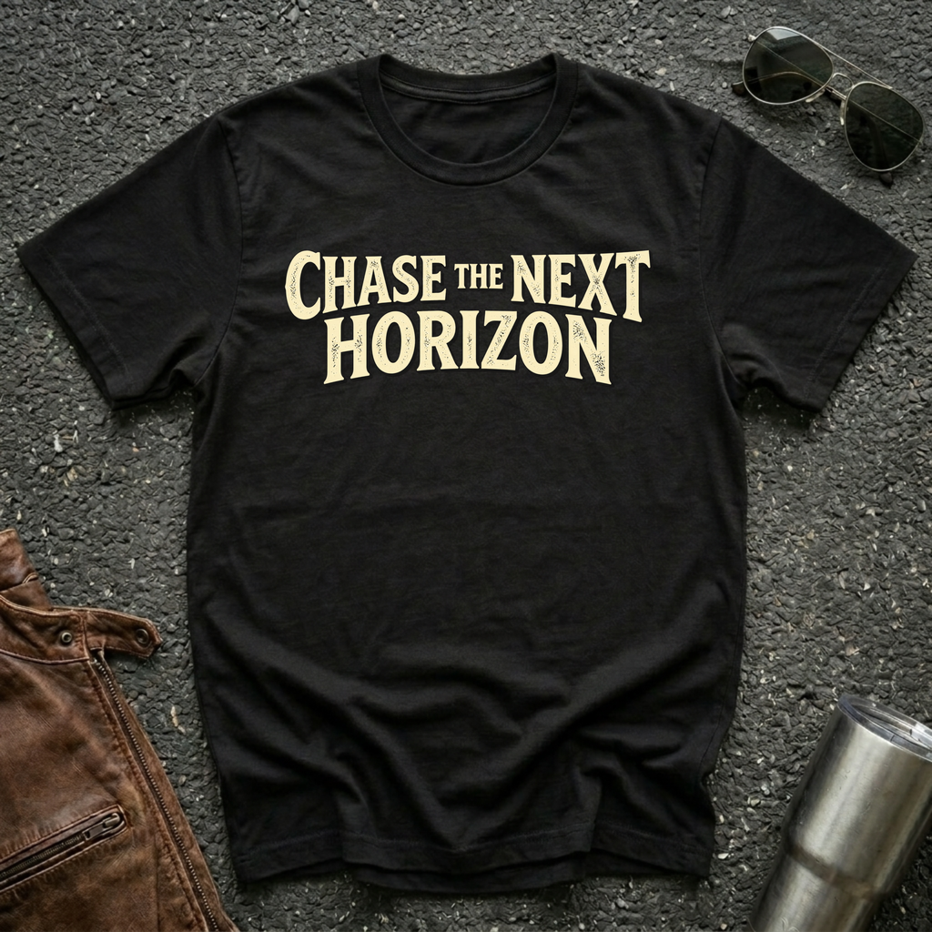A black short-sleeve t-shirt with the phrase 'CHASE THE NEXT HORIZON' printed in a distressed beige font across the chest, laid flat on a dark asphalt-like surface with sunglasses, a brown leather jacket, and a metal tumbler nearby.