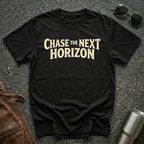 A black short-sleeve t-shirt with the phrase 'CHASE THE NEXT HORIZON' printed in a distressed beige font across the chest, laid flat on a dark asphalt-like surface with sunglasses, a brown leather jacket, and a metal tumbler nearby.