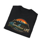A black folded t-shirt with a retro-style scenic graphic on the front. The graphic features mountains, a winding river, pine trees, and an orange sunset, with the text 'The Riverstone Life' and 'Fueled by Freedom'.