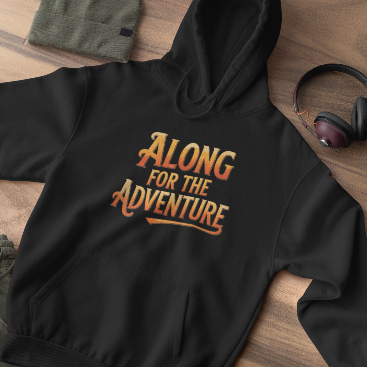 A black pullover hoodie laid flat on a wooden surface, featuring the phrase 'ALONG FOR THE ADVENTURE' printed across the chest in a stylized orange-to-yellow gradient. A folded olive green beanie, a pair of headphones, and a small shoe are partially visible as styled props.
