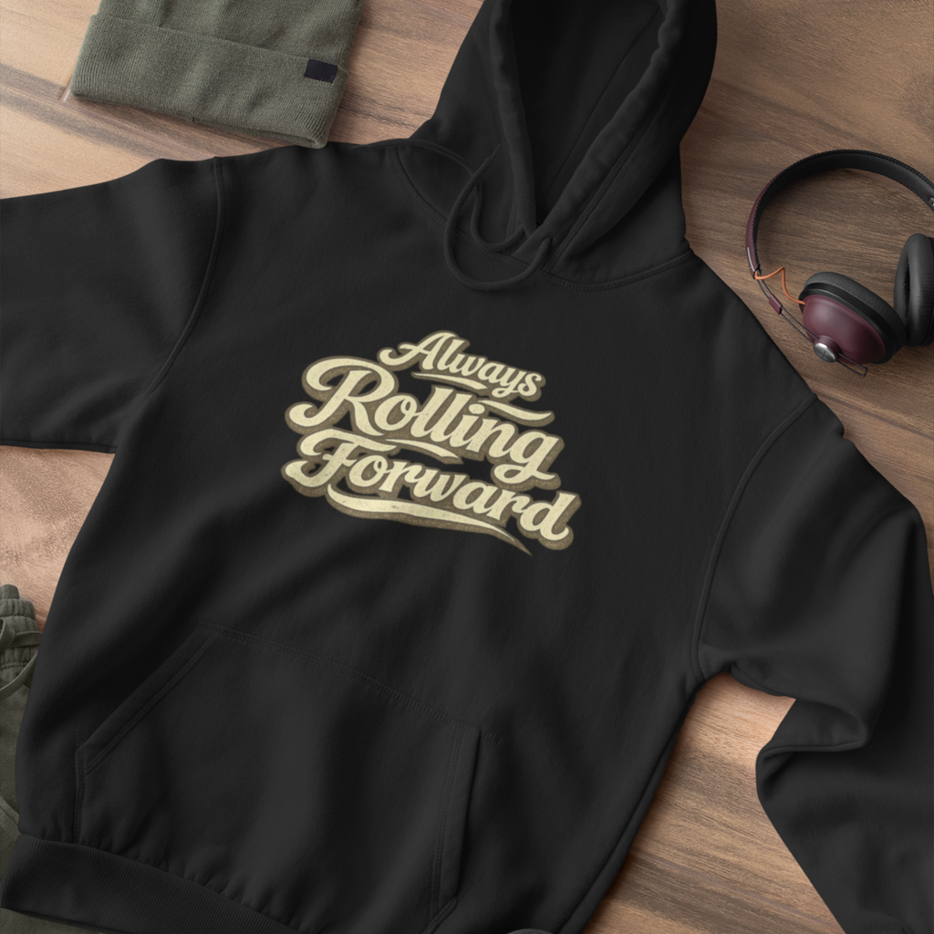 A black pullover hoodie laid flat on a wooden surface, featuring the phrase 'Always Rolling Forward' printed across the chest in a beige script font. A folded olive green garment, a pair of over-ear headphones, and a small dark accessory are placed around it as styling props.