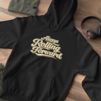 A black pullover hoodie laid flat on a wooden surface, featuring the phrase 'Always Rolling Forward' printed across the chest in a beige script font. A folded olive green garment, a pair of over-ear headphones, and a small dark accessory are placed around it as styling props.