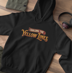 A black pullover hoodie laid flat on a wooden surface, featuring the phrase 'Chasing the Yellow Lines' in a stylized yellow and red graphic across the chest. A folded olive green beanie, a pair of black over-ear headphones, and a small portion of another garment are placed around it as styling props.