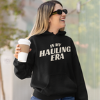 A woman wearing a black hoodie with the text 'IN MY HAULING ERA' in white, holding a coffee cup outdoors.