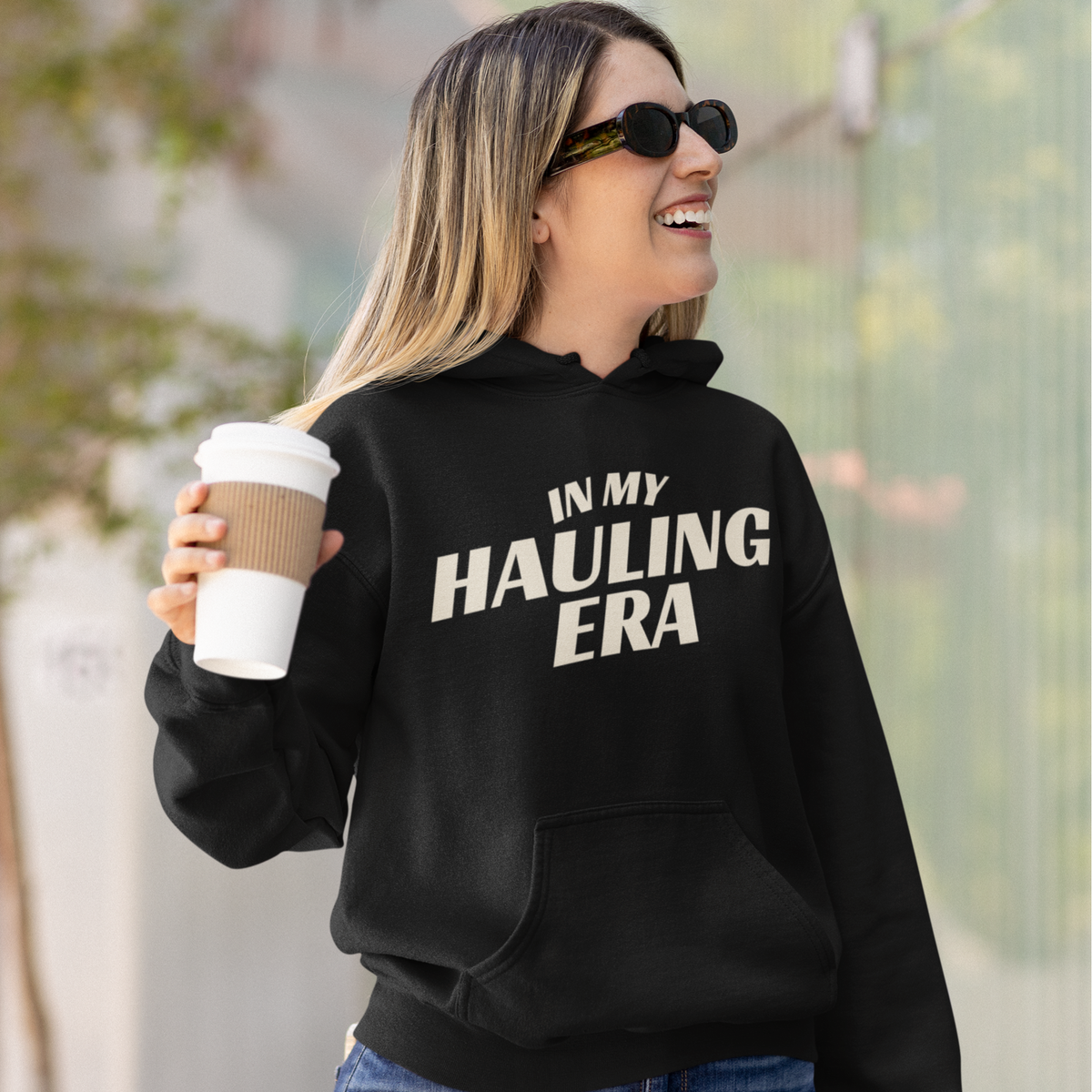 A woman wearing a black hoodie with the text 'IN MY HAULING ERA' in white, holding a coffee cup outdoors.