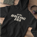 A black pullover hoodie laid flat on a wooden surface, featuring the phrase 'IN MY HAULING ERA' printed in large white letters across the chest. A folded olive green fabric item and a pair of over-ear headphones are partially visible as styled props.