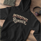 A black pullover hoodie laid flat on a wooden surface, featuring the words 'INTERSTATE RUNNER' in a stylized cream and brown script across the chest. A folded olive green garment, a pair of headphones, and a small dark accessory are placed around it as styling props.