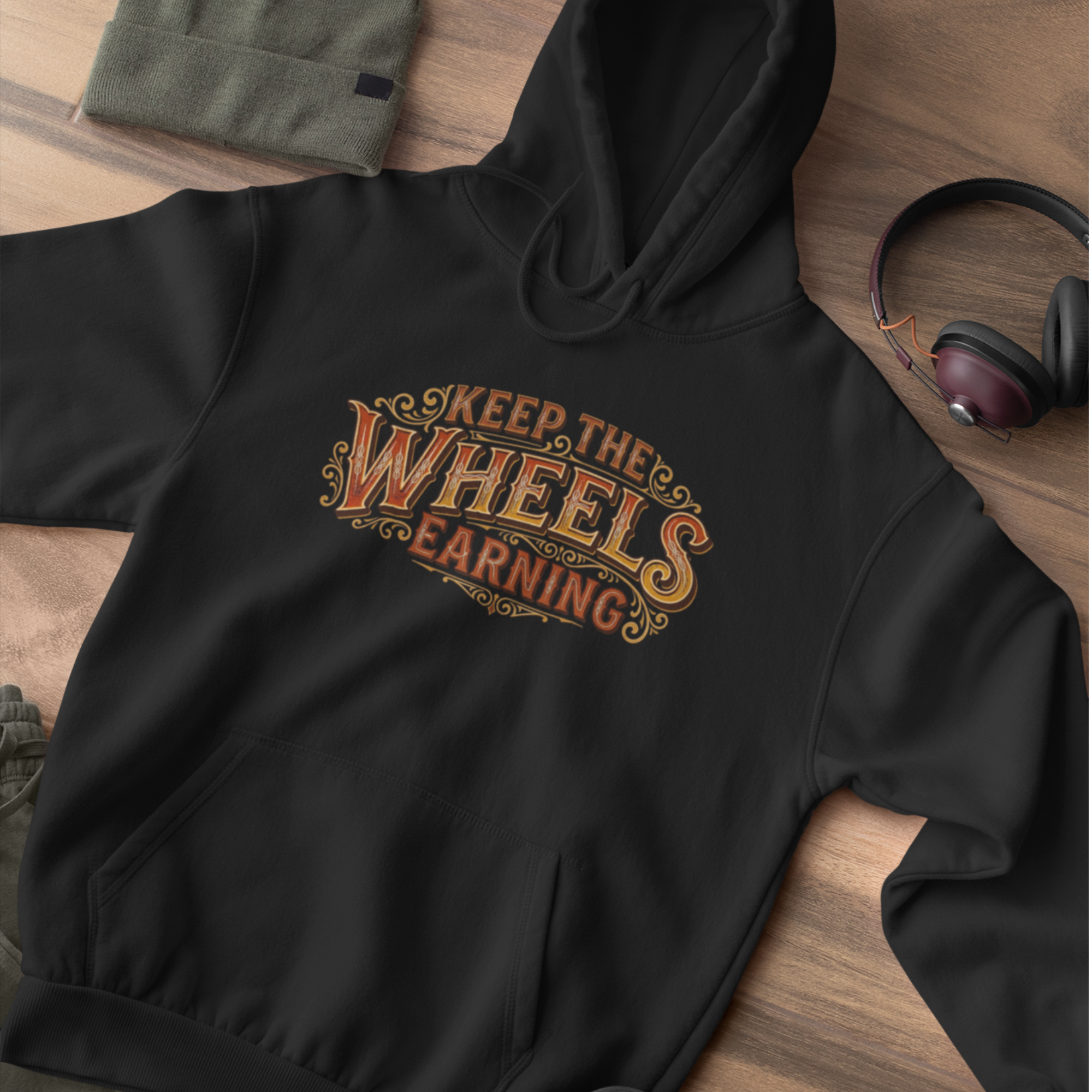 A black pullover hoodie laid flat on a wooden surface, featuring the phrase 'KEEP THE WHEELS EARNING' in a decorative orange and gold graphic across the chest. A folded olive green garment, a pair of headphones, and a small fabric item are placed around it as styling props.