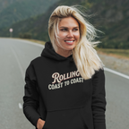 A smiling blonde woman stands on a road with mountains in the background, wearing a black hoodie with the words 'Rolling Coast to Coast' printed across the front in a light beige script and block font.