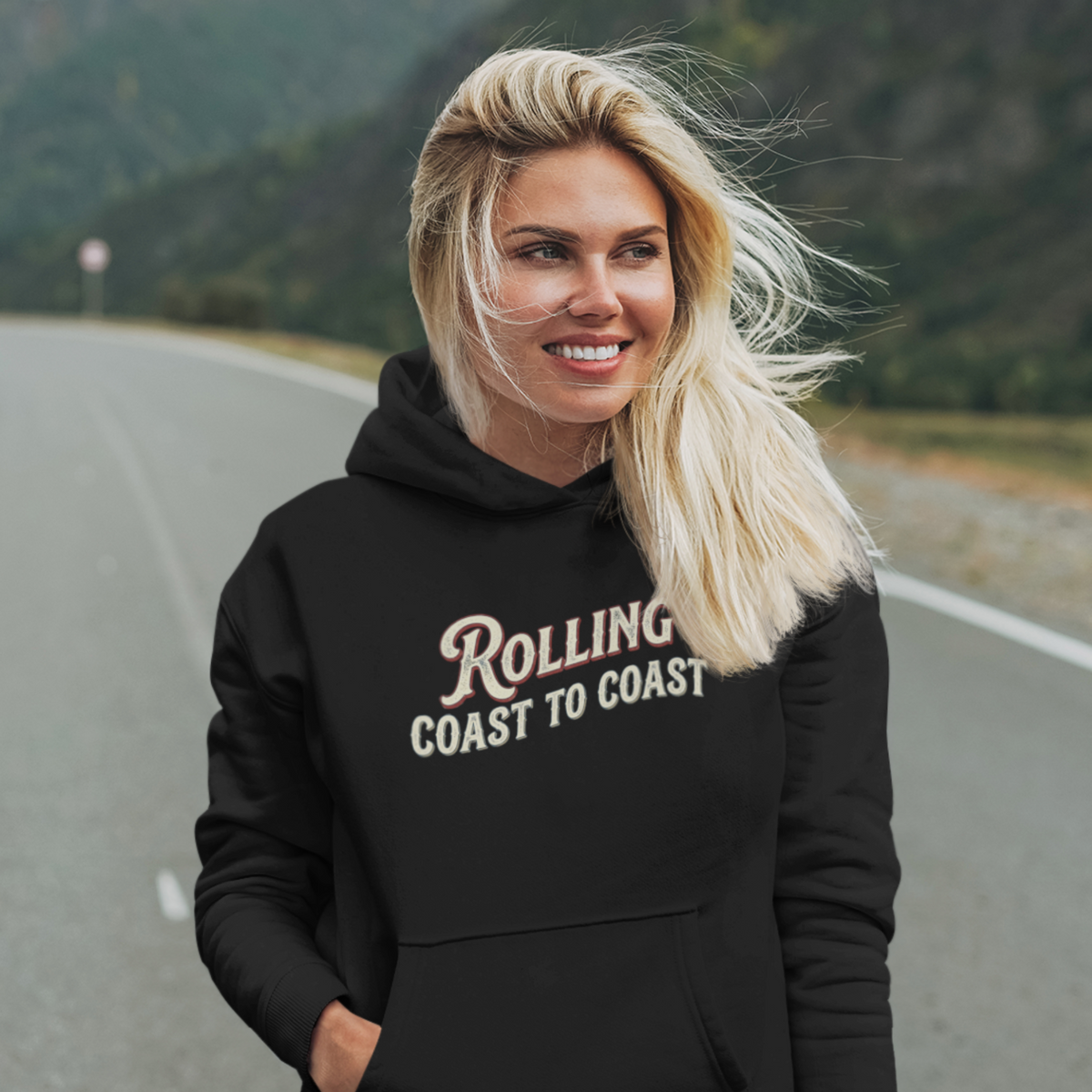 A smiling blonde woman stands on a road with mountains in the background, wearing a black hoodie with the words 'Rolling Coast to Coast' printed across the front in a light beige script and block font.