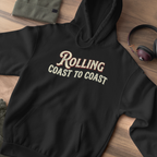 A black pullover hoodie laid flat on a wood surface, featuring the phrase 'Rolling Coast to Coast' printed across the chest in a cream and burgundy retro-style font. A folded olive green sweatshirt, a pair of over-ear headphones, and part of another garment are visible around it as styling props.