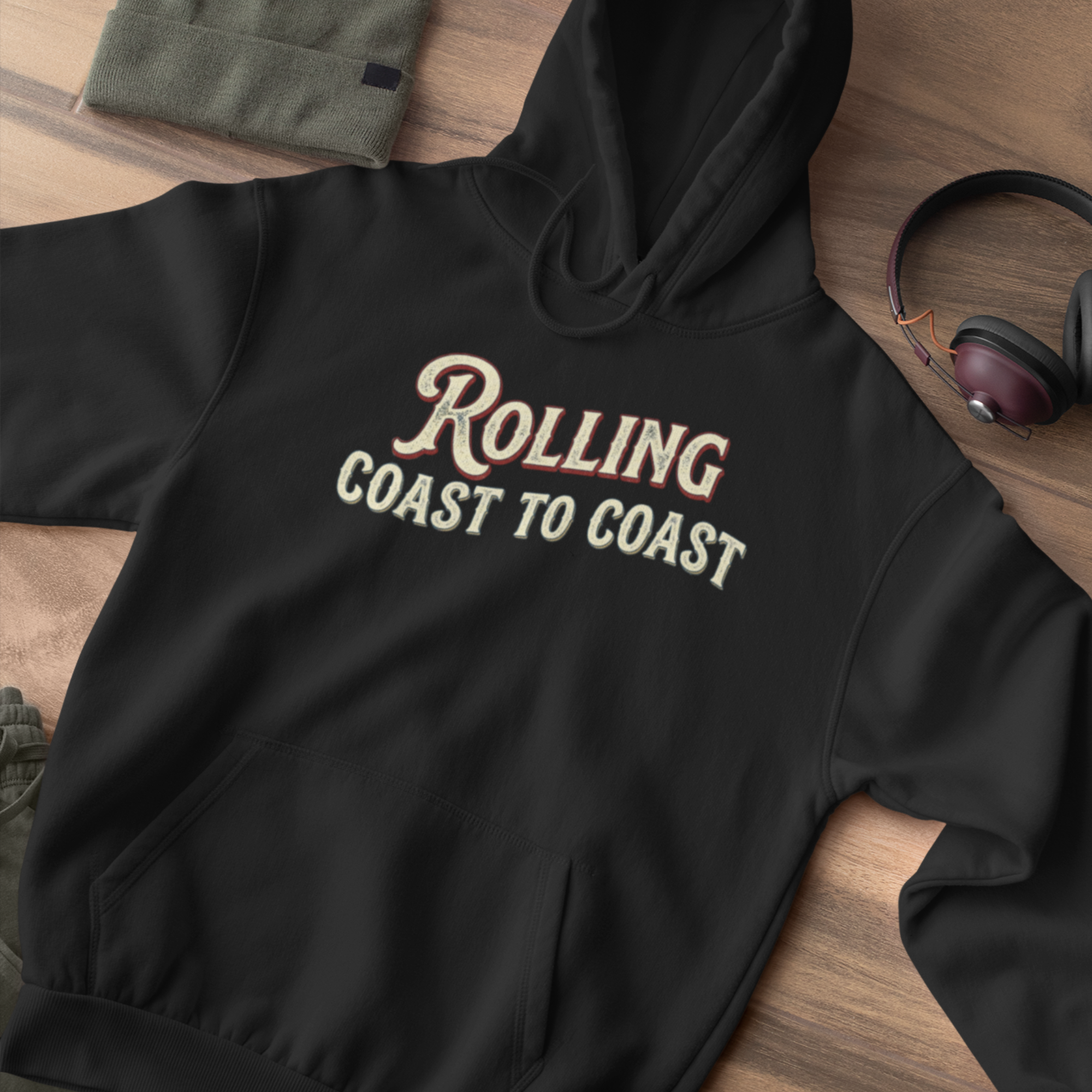 A black pullover hoodie laid flat on a wood surface, featuring the phrase 'Rolling Coast to Coast' printed across the chest in a cream and burgundy retro-style font. A folded olive green sweatshirt, a pair of over-ear headphones, and part of another garment are visible around it as styling props.