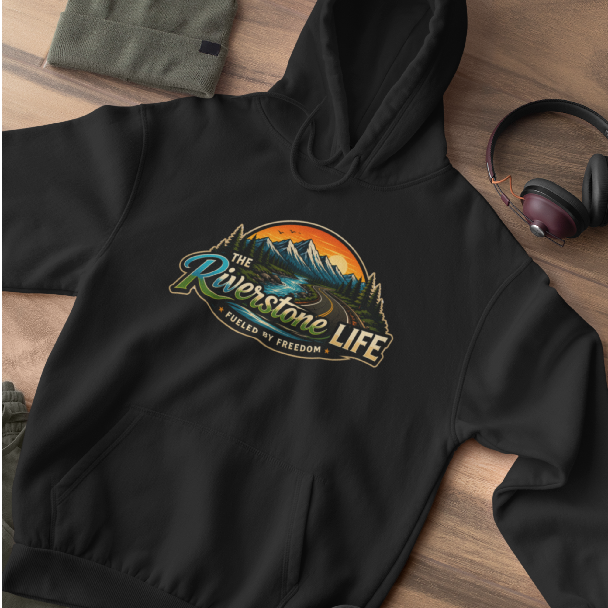 A black pullover hoodie laid flat on a wooden surface, featuring a large retro-style mountain landscape chest graphic with the text 'The Riverstone Life' and 'Fueled by Freedom.' A folded olive green fabric item and over-ear headphones are partially visible as styling props.