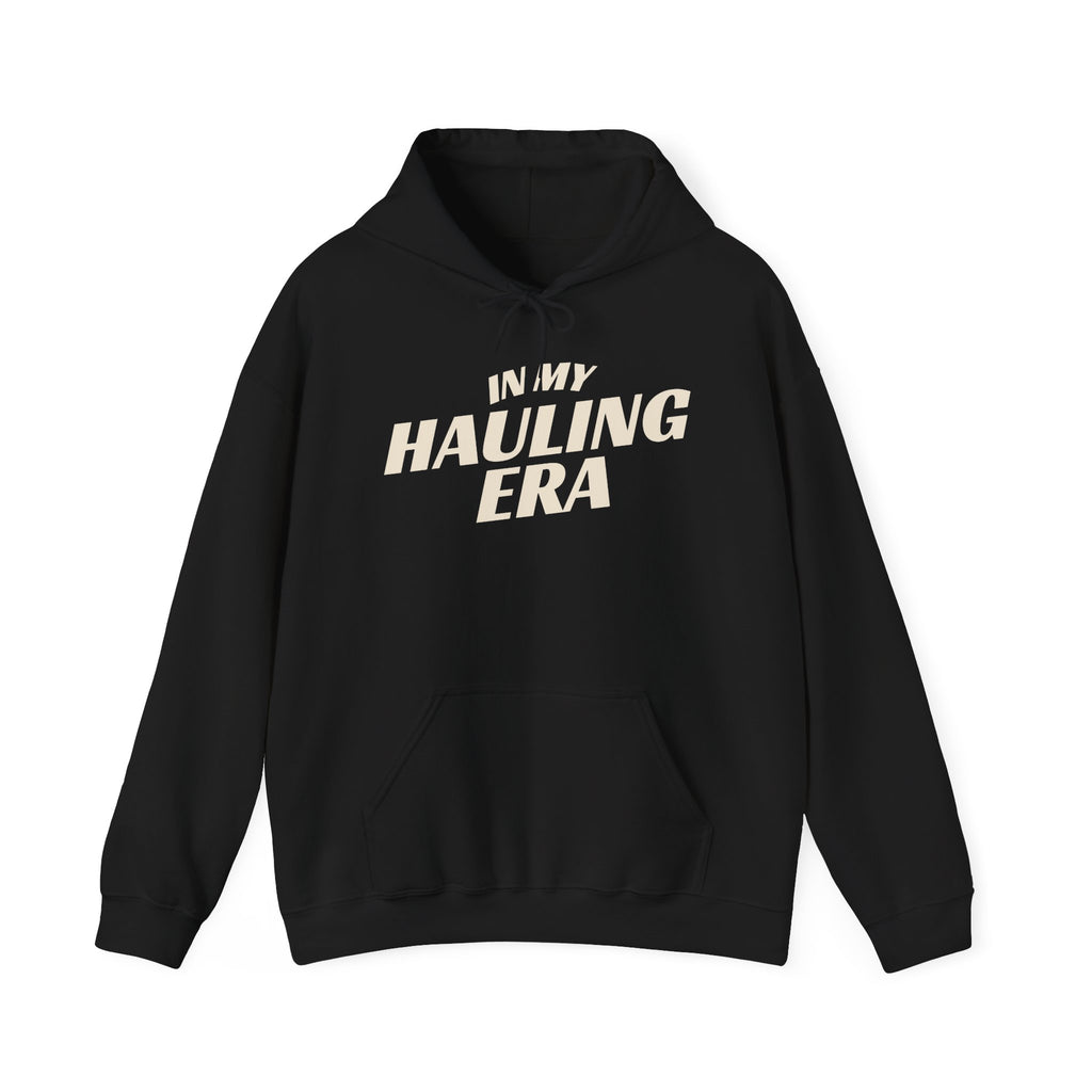 A black pullover hoodie with the phrase 'IN MY HAULING ERA' printed in a bold, cream-colored font across the chest, displayed on a clean white background.