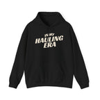 A black pullover hoodie with the phrase 'IN MY HAULING ERA' printed in a bold, cream-colored font across the chest, displayed on a clean white background.
