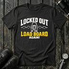A black graphic t-shirt laid flat on a dark wood background with boots, a bottle, and a gauge as props. The shirt features a graphic that reads 'LOCKED OUT OF THE LOAD BOARD AGAIN!' in white and yellow text with a padlock and chain illustration.