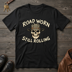 A black graphic t-shirt featuring a skull wearing a cowboy hat with the text 'ROAD WORN STILL ROLLING' in a distressed cream font. The t-shirt is displayed in a flat-lay arrangement on a dark wood background, surrounded by decorative props including keys, leather shoes, and a brown leather jacket.