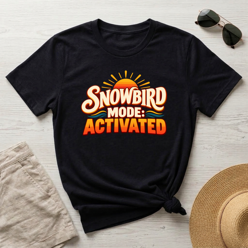 A black short-sleeve graphic T-shirt laid flat on a light wood surface, styled with sunglasses, beige shorts, and a straw hat. The shirt has a large front graphic reading “SNOWBIRD MODE: ACTIVATED” in orange, cream, and yellow tones with a sun and small blue wave accents.