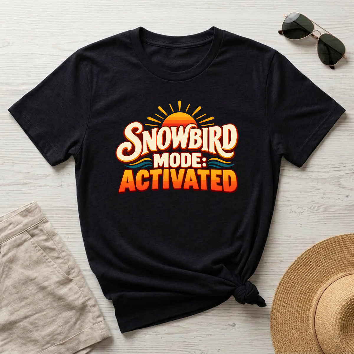 A black short-sleeve graphic T-shirt laid flat on a light wood surface, styled with sunglasses, beige shorts, and a straw hat. The shirt has a large front graphic reading “SNOWBIRD MODE: ACTIVATED” in orange, cream, and yellow tones with a sun and small blue wave accents.