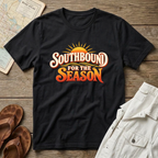 A black short-sleeve graphic T-shirt laid flat on a rustic wood surface, with a retro-style sun graphic and the words “SOUTHBOUND FOR THE SEASON” in white and orange. Surrounding props include a map, brown sandals, and light-colored shorts.