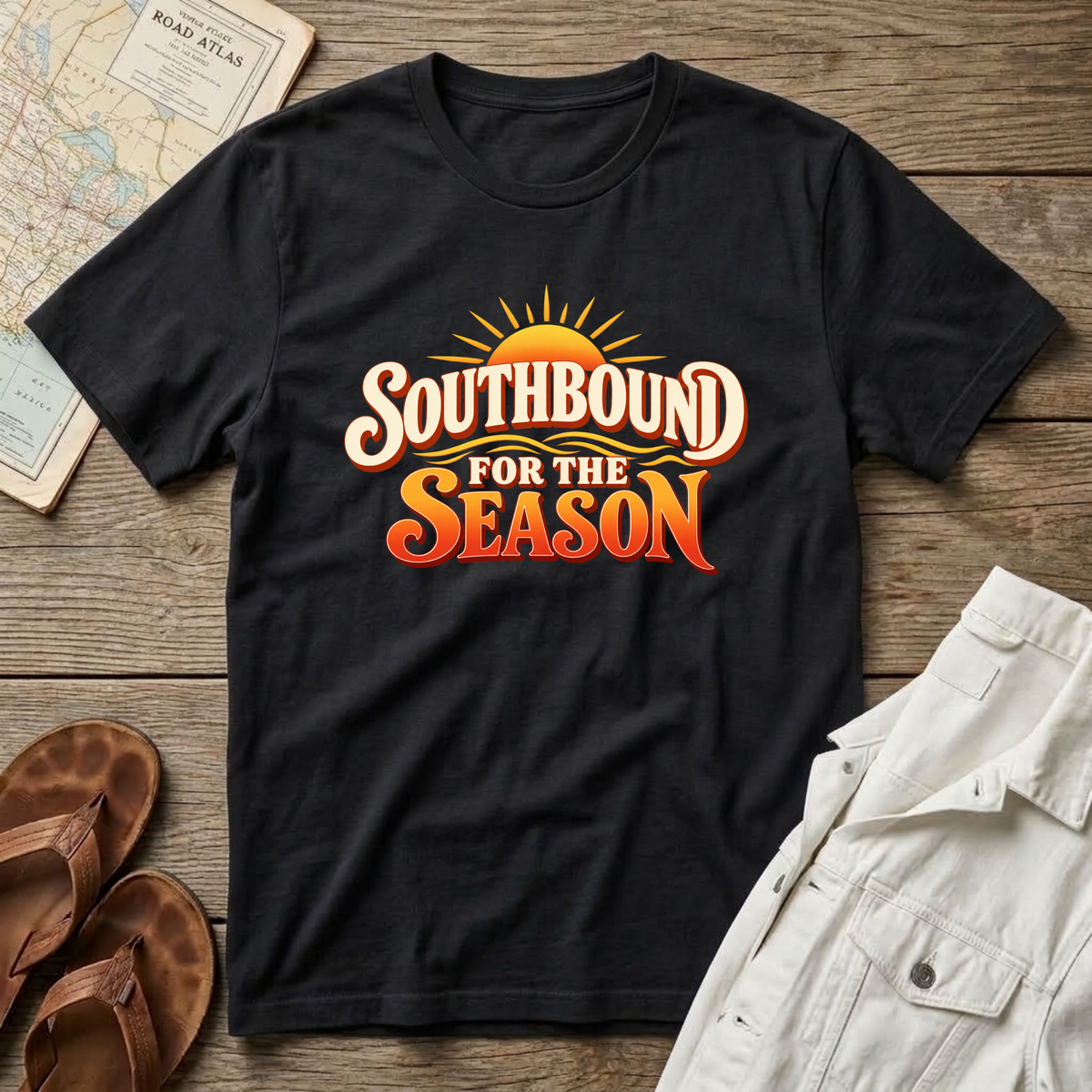 A black short-sleeve graphic T-shirt laid flat on a rustic wood surface, with a retro-style sun graphic and the words “SOUTHBOUND FOR THE SEASON” in white and orange. Surrounding props include a map, brown sandals, and light-colored shorts.