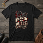 A black graphic t-shirt featuring a raven and the text 'EARNED MY MILES' with 'NOTHING LEFT TO PROVE' on a distressed banner. The shirt is displayed in a flat-lay arrangement on a dark wood background, accompanied by keys, brown leather shoes, and a brown leather jacket.