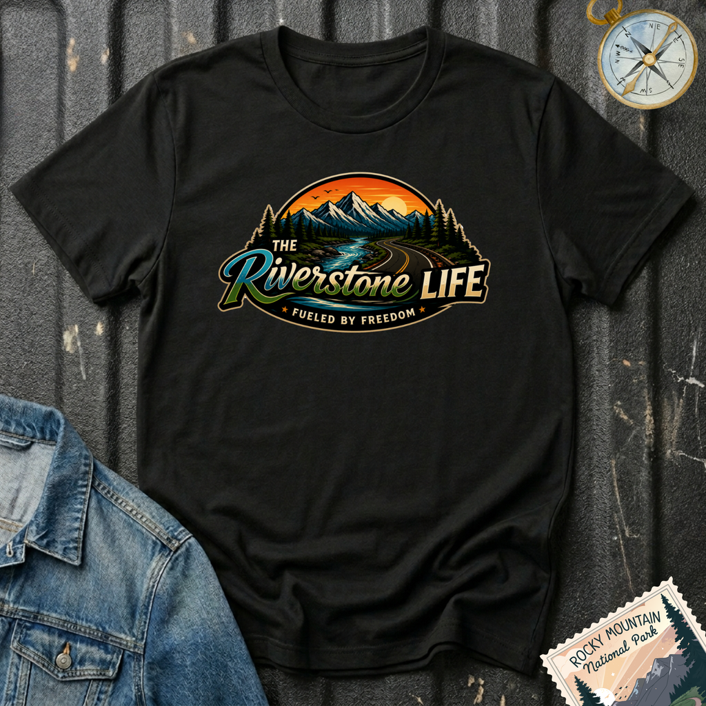 A black short-sleeve graphic T-shirt laid flat on a dark textured surface, featuring a scenic mountain and river illustration with the text 'The Riverstone Life' and 'Fueled by Freedom.' A denim jacket corner, a compass, and a Rocky Mountain National Park stamp are placed around it as styling props.