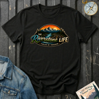 A black short-sleeve graphic T-shirt laid flat on a dark textured surface, featuring a scenic mountain and river illustration with the text 'The Riverstone Life' and 'Fueled by Freedom.' A denim jacket corner, a compass, and a Rocky Mountain National Park stamp are placed around it as styling props.