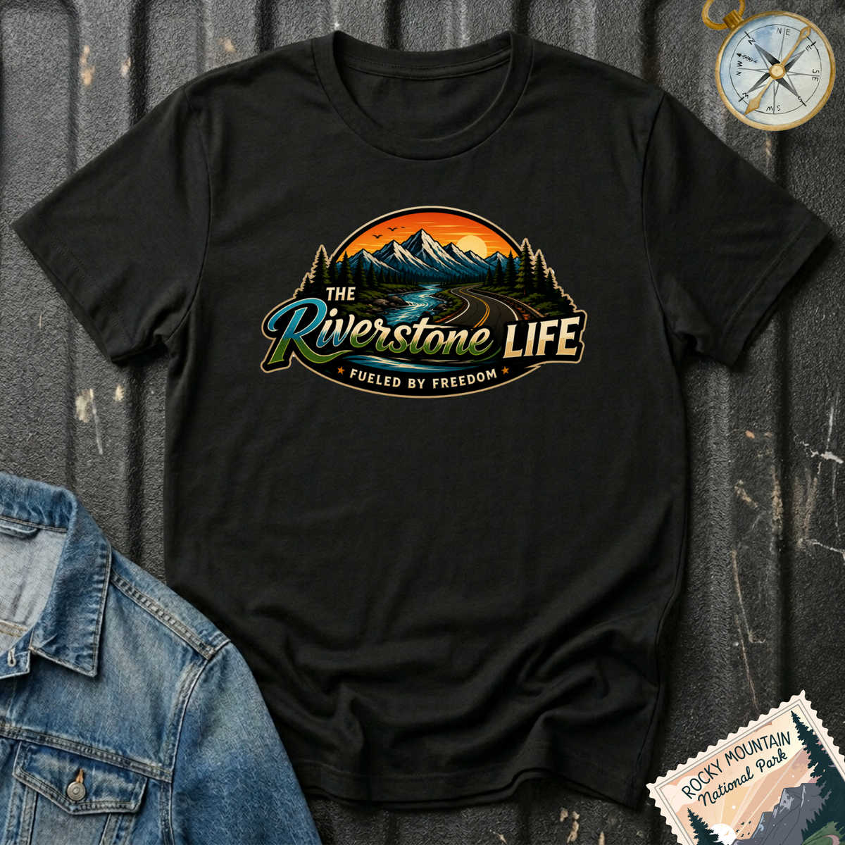 A black short-sleeve graphic T-shirt laid flat on a dark textured surface, featuring a scenic mountain and river illustration with the text 'The Riverstone Life' and 'Fueled by Freedom.' A denim jacket corner, a compass, and a Rocky Mountain National Park stamp are placed around it as styling props.
