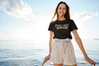 A woman stands on a boat wearing a black graphic t-shirt with the text 'CHASE THE NEXT HORIZON' in gold, paired with cream and navy pinstriped shorts, against a bright ocean and sky background.