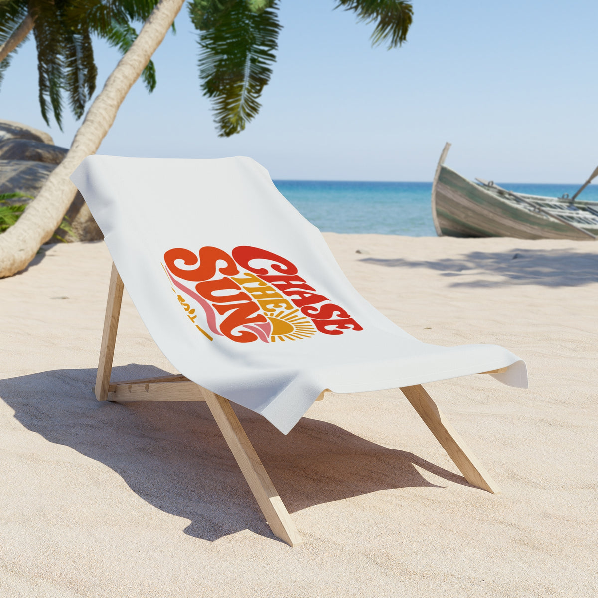 A white beach towel draped over a wooden lounge chair on a sandy beach, featuring a retro-style red and orange graphic that reads 'Chase the Sun'.