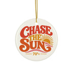 A round ceramic Christmas ornament with a white glossy background, featuring retro-style text that reads 'CHASE THE SUN' in red and orange, with a yellow sun graphic and '70+' below. The ornament hangs from a gold string against a plain white background.