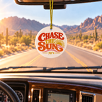 A round white ornament hanging from a yellow string in a car, featuring the text 'Chase the Sun 70+' in red and orange lettering with a yellow sun graphic. The background shows a scenic desert road with mountains and cacti seen through the windshield.