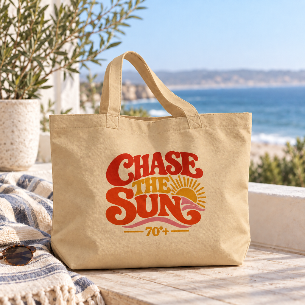 A beige canvas tote bag with a retro-style 'Chase the Sun 70+' graphic, displayed on a wooden deck with a beach and ocean background.