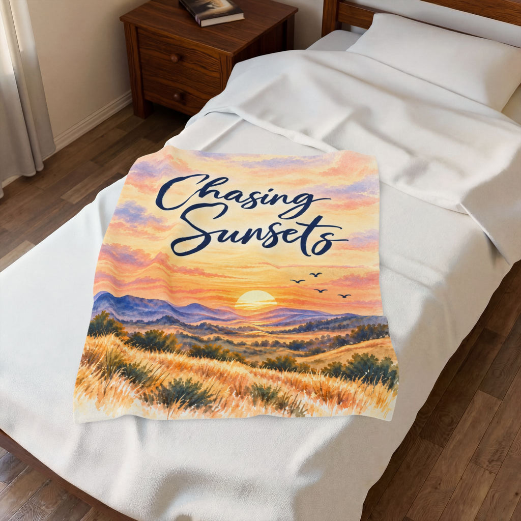 A white fleece blanket with a colorful sunset landscape print and the text 'Chasing Sunsets' in navy script, displayed on a neatly made bed in a bedroom setting.