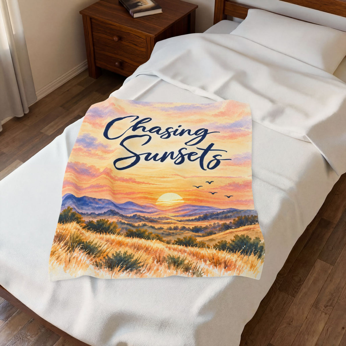 A white fleece blanket with a colorful sunset landscape print and the text 'Chasing Sunsets' in navy script, displayed on a neatly made bed in a bedroom setting.