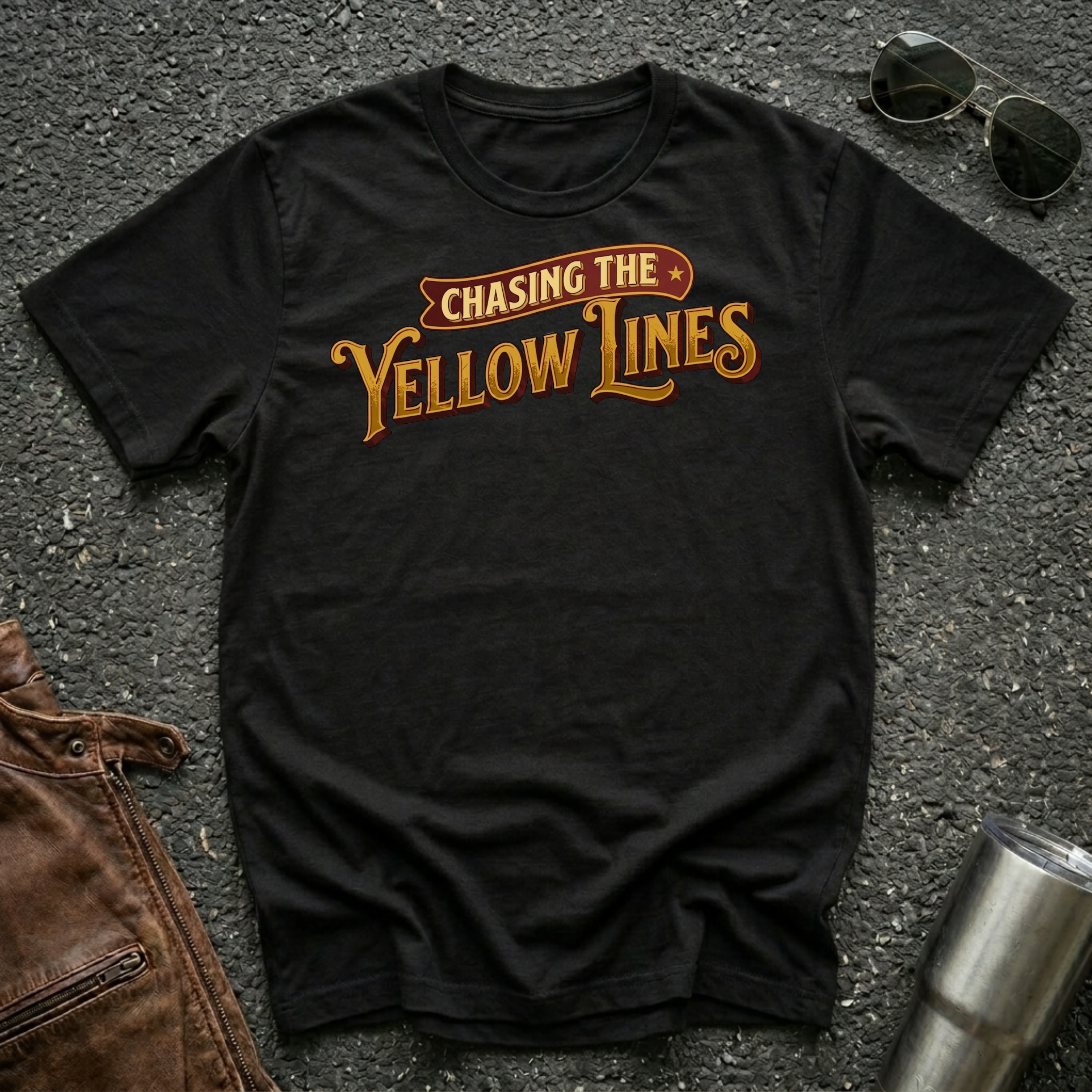 A black short-sleeve graphic T-shirt laid flat on a dark textured surface, with sunglasses, brown leather jacket, and a metal tumbler nearby. The shirt has a large retro-style chest print reading “Chasing The Yellow Lines” in orange and yellow tones.