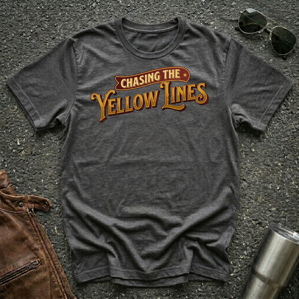 A dark heather short-sleeve graphic T-shirt laid flat on a dark textured surface, with sunglasses, brown leather jacket, and a metal tumbler nearby. The shirt has a large retro-style chest print reading “Chasing The Yellow Lines” in orange and yellow tones.