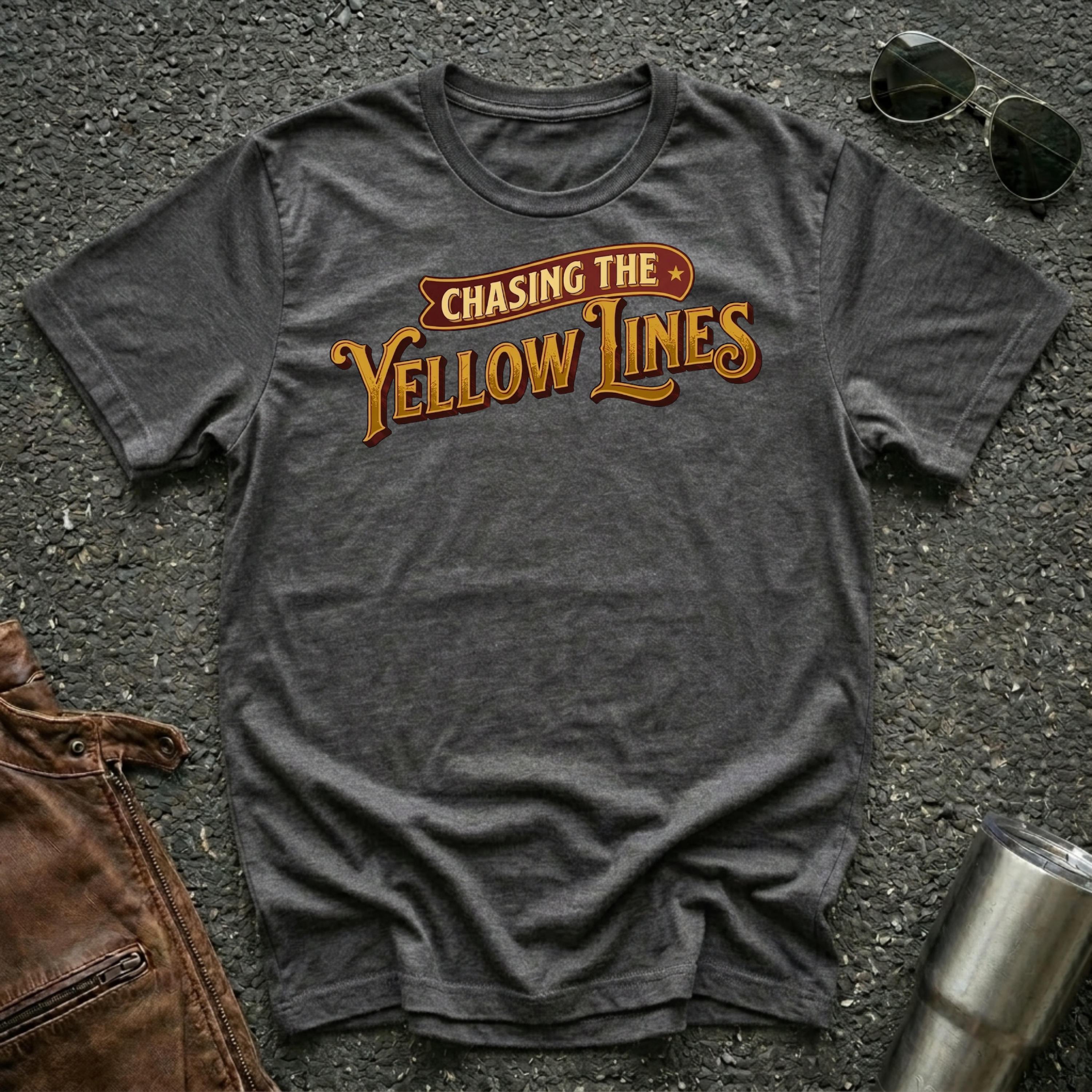 A dark heather short-sleeve graphic T-shirt laid flat on a dark textured surface, with sunglasses, brown leather jacket, and a metal tumbler nearby. The shirt has a large retro-style chest print reading “Chasing The Yellow Lines” in orange and yellow tones.