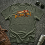 A military green short-sleeve graphic T-shirt laid flat on a dark textured surface, with sunglasses, brown leather jacket, and a metal tumbler nearby. The shirt has a large retro-style chest print reading “Chasing The Yellow Lines” in orange and yellow tones.