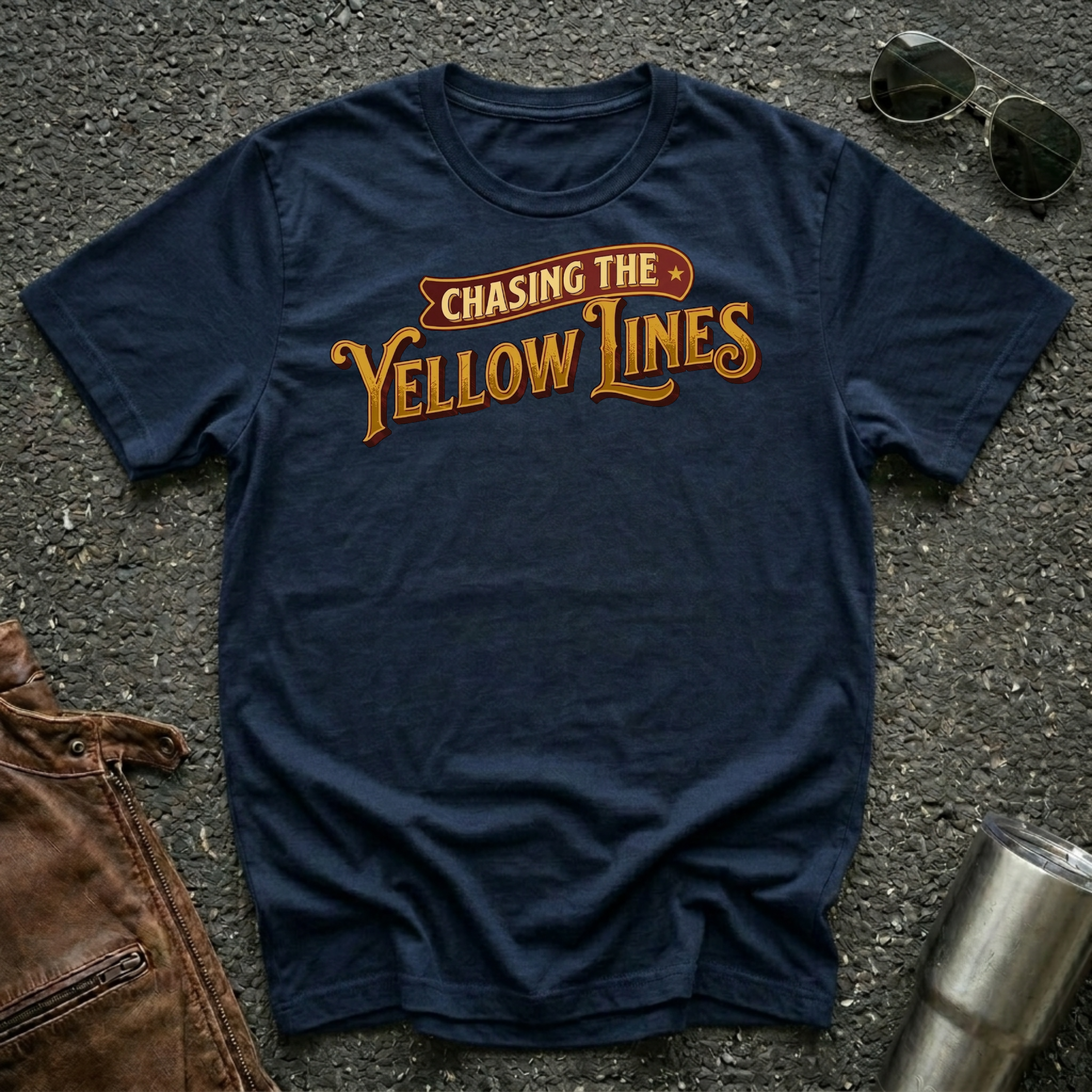 A navy short-sleeve graphic T-shirt laid flat on a dark textured surface, with sunglasses, brown leather jacket, and a metal tumbler nearby. The shirt has a large retro-style chest print reading “Chasing The Yellow Lines” in orange and yellow tones.