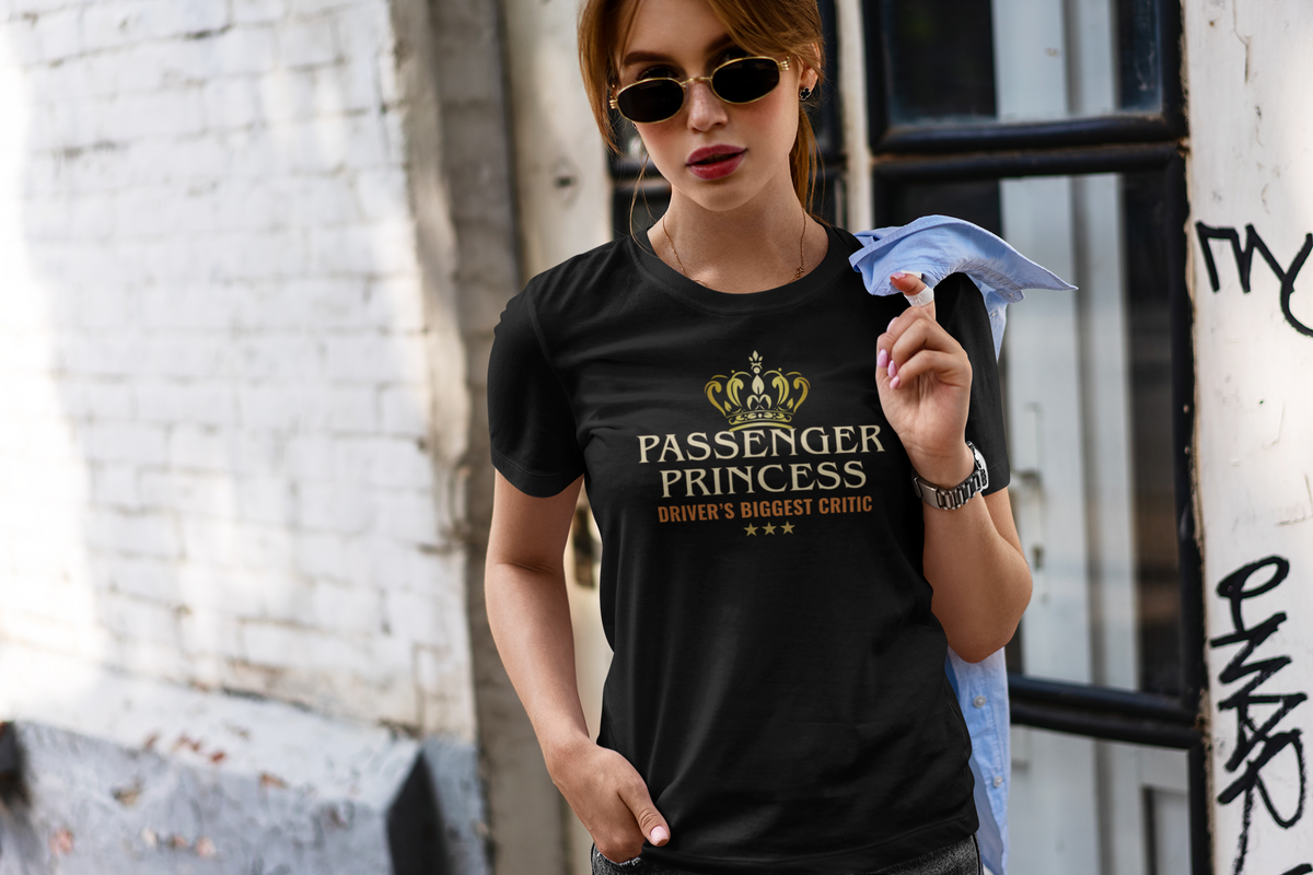A woman wearing a black graphic t-shirt with a gold crown and the text 'PASSENGER PRINCESS DRIVER'S BIGGEST CRITIC' stands in an urban setting.