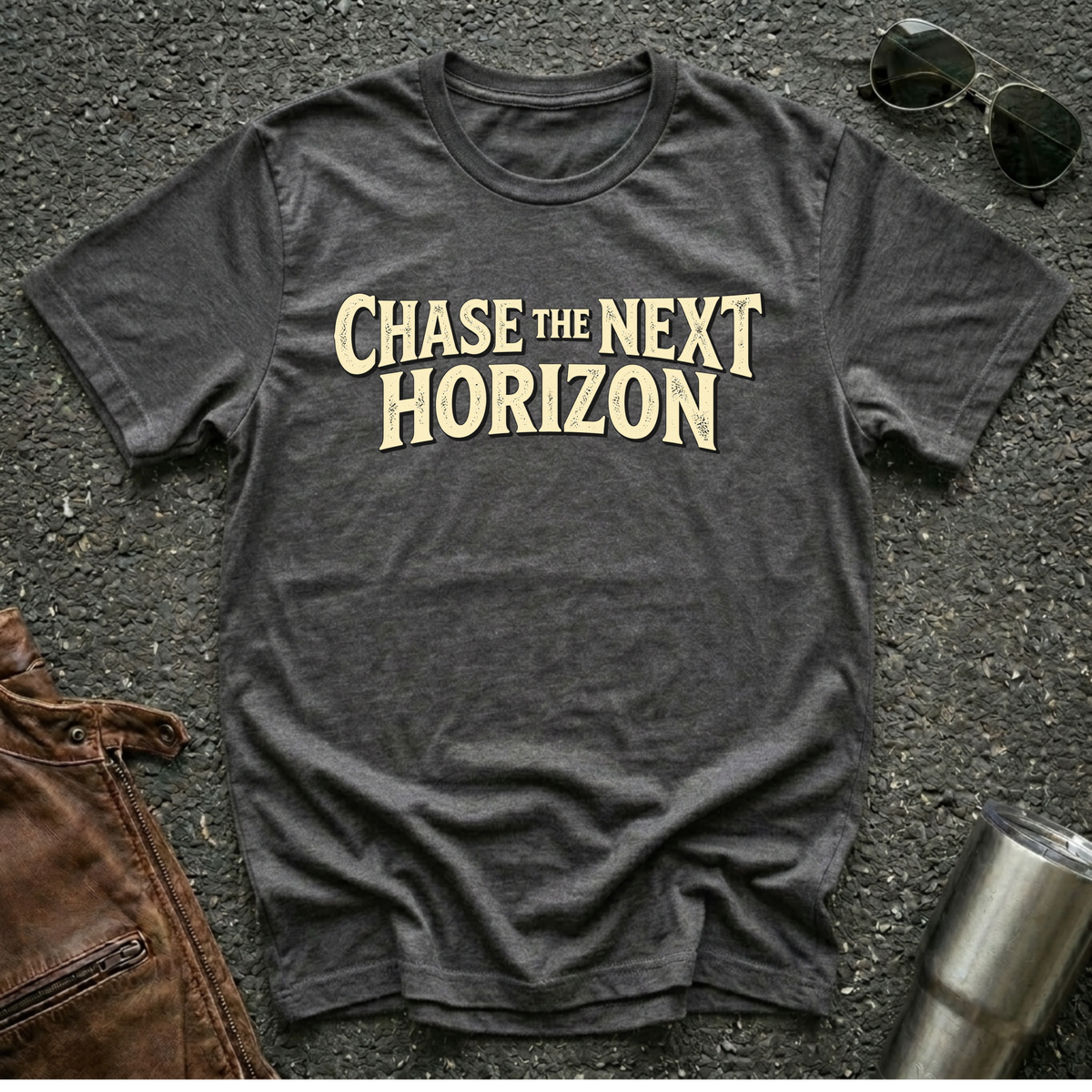A dark heather gray t-shirt with the phrase 'CHASE THE NEXT HORIZON' printed in a distressed cream-colored font across the chest. The shirt is presented in a flat-lay arrangement on a textured asphalt background, styled with sunglasses, a brown leather jacket, and a stainless steel tumbler.