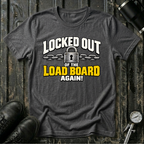 A dark heather gray graphic t-shirt laid flat on a rustic dark wood background, styled with black leather boots  and a tire gauge. The t-shirt features a graphic print that reads 'LOCKED OUT OF THE LOAD BOARD AGAIN!' in white and yellow lettering with a padlock and chain illustration.