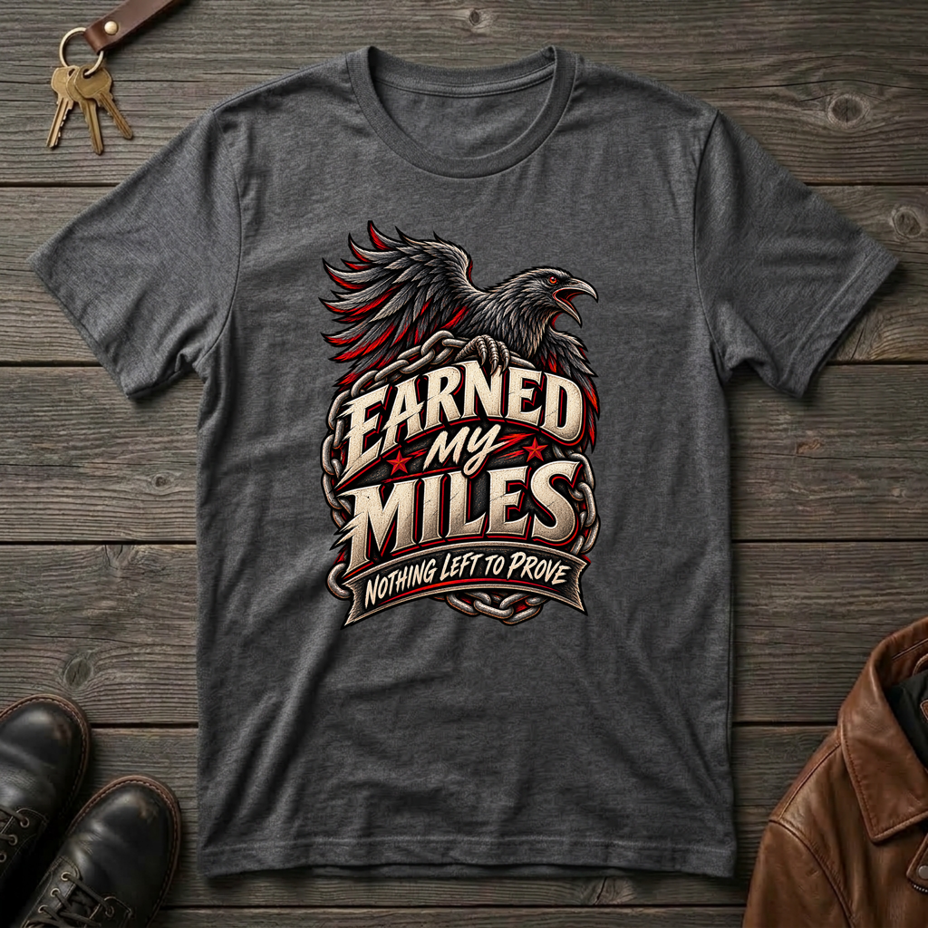 A dark heather gray graphic t-shirt featuring a raven and the text 'Earned My Miles Nothing Left to Prove', laid flat on a rustic dark wood background with keys, shoes, and a leather jacket as props.