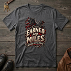 A dark heather gray graphic t-shirt featuring a raven and the text 'Earned My Miles Nothing Left to Prove', laid flat on a rustic dark wood background with keys, shoes, and a leather jacket as props.